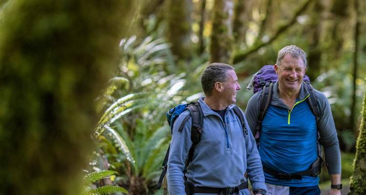 Deux randonneurs masculins souriants marchant dans une forêt mousseuse luxuriante tout en portant des sacs à dos de jour.
