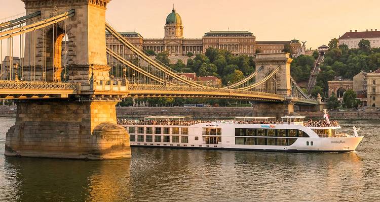 Barco de crucero fluvial pasando bajo el Puente de las Cadenas de Budapest al atardecer con el Castillo de Buda de fondo.
