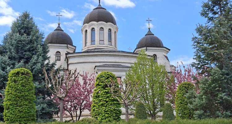 Iglesia blanca y elegante con tres cúpulas oscuras enmarcadas por árboles en flor y arbustos bien cuidados.