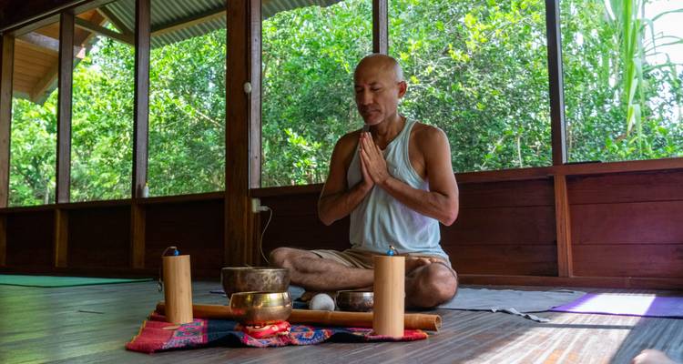 Un homme assis en position du lotus dans un studio en bois en plein air effectuant un rituel de méditation avec des bols chantants.
