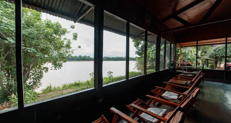 Überdachtes Flussuferdeck mit Liegestühlen aus Holz, die einem breiten schlammigen Fluss durch Siebfenster zugewandt sind.