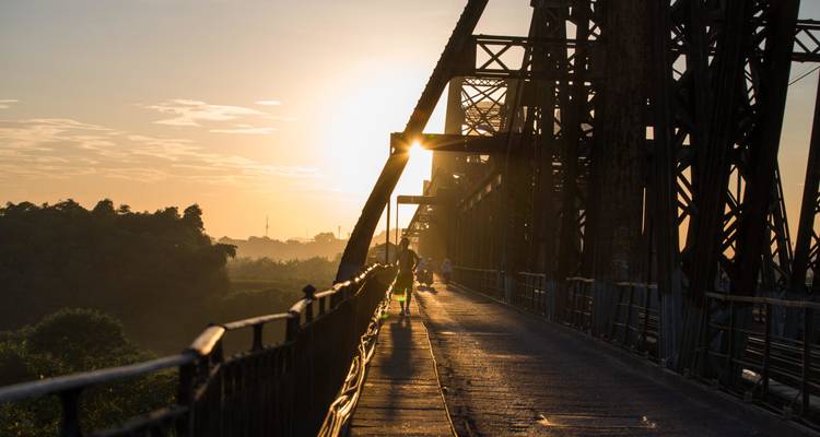 Le soleil du matin brille à travers les poutrelles d'un long pont en fer tandis que quelques piétons et cyclistes le traversent.