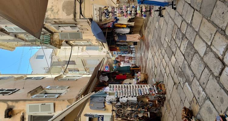 Ruelle étroite pavée de galets dans la vieille ville de Corfou, bordée de petits commerces de souvenirs et de visiteurs en promenade.