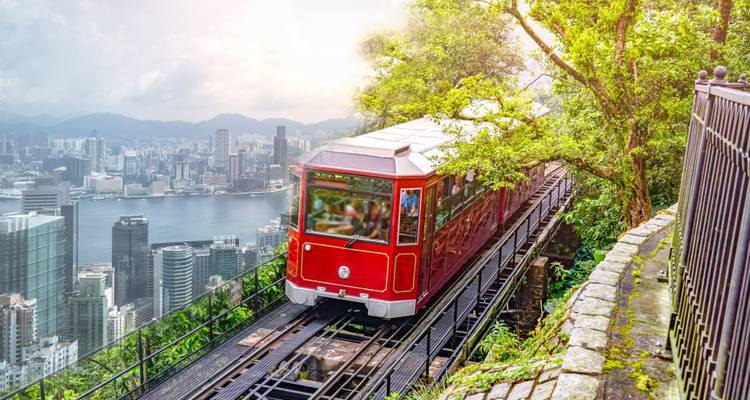 Historische rote Peak Tram, die eine steile Strecke durch üppige Vegetation erklimmt, mit panoramischer Skyline von Hongkong im Hintergrund.