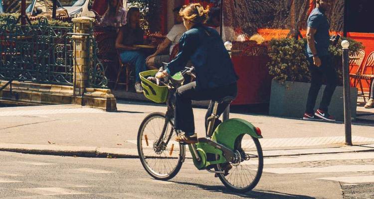 Ciclista en una bici verde pasando por los asientos de una cafetería al aire libre bajo el sol brillante.