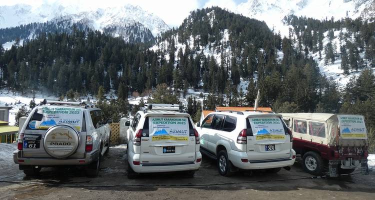 Line of expedition SUVs with mountain banners parked at snow-covered forest base camp.