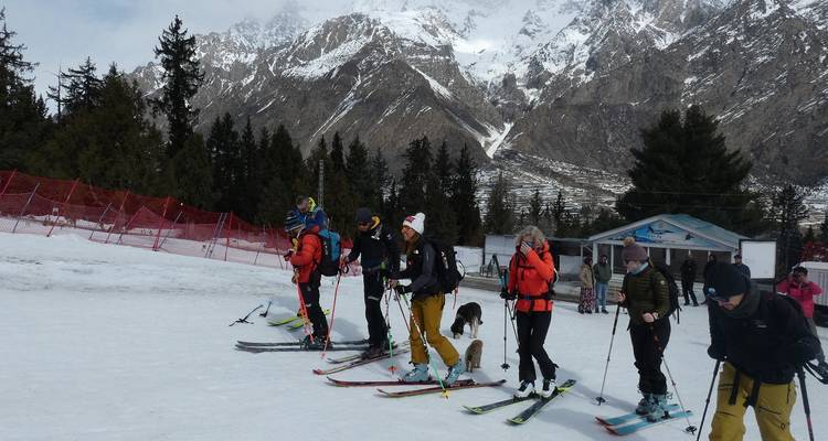 Row of skiers lined up on snow slope with forest and alpine ridge in background.