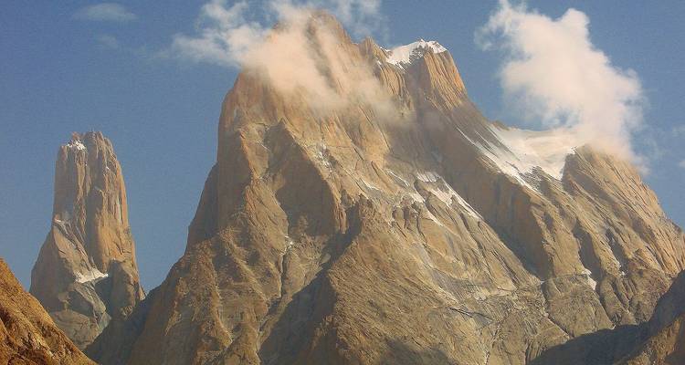 Steep granite spires of Trango Towers partially shrouded in clouds against blue sky.