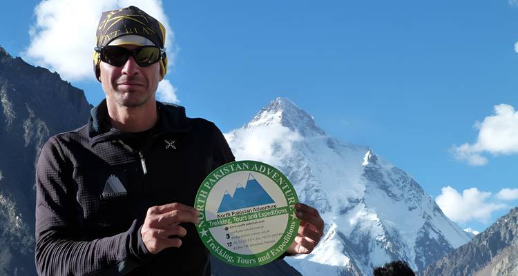 Trekker holding expedition logo plate with snow-covered K2 massif in background.