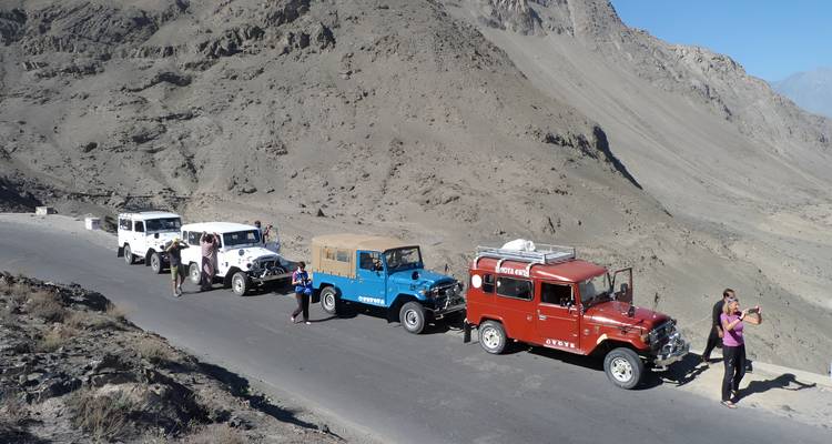 Convoy of colorful jeeps parked along mountain road with passengers taking photos.