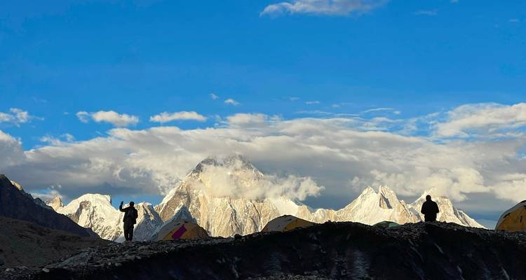 Silhouettes of trekkers and yellow tents with towering snow-covered Karakoram peaks glowing in evening light.