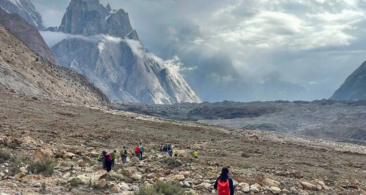 Small trekking group crossing a rocky glacier valley with dramatic jagged peaks and clouds ahead.