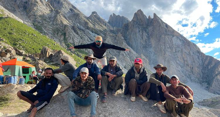 Trekking team resting on a boulder with steep rocky spires towering behind them under a partly cloudy sky.