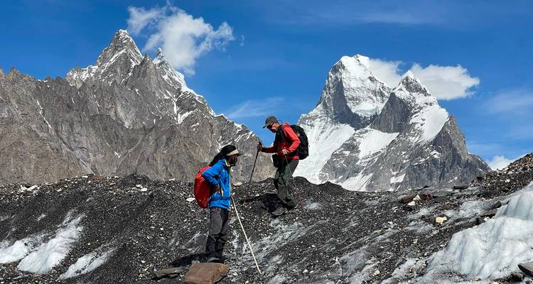 Two climbers with backpacks ascend black moraine rubble with snow-capped Karakoram peaks rising behind.