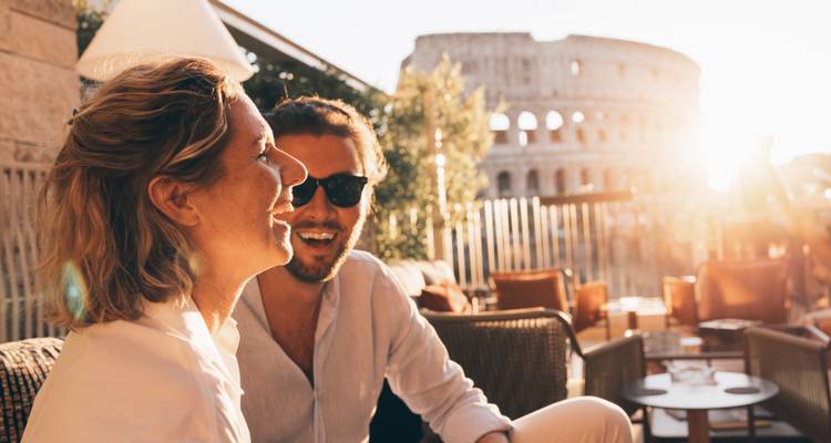 Un couple rit en prenant des verres sur une terrasse avec le Colosseum doucement illuminé par le coucher de soleil en arrière-plan.