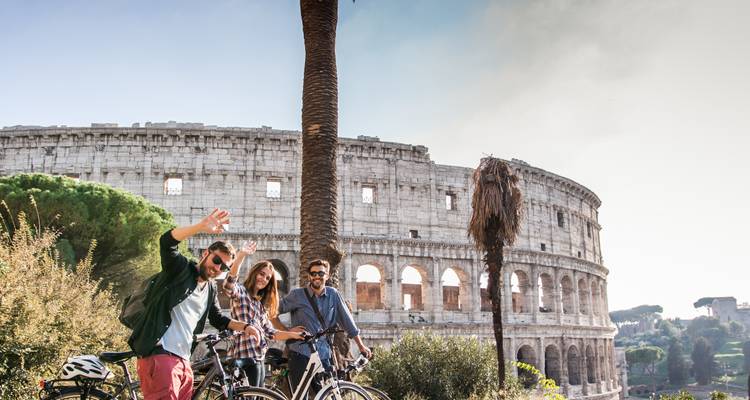 Drie vrienden met fietsen die poseren en zwaaien voor het Colosseum van Rome op een helder dag.