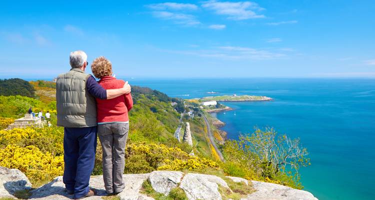 Un couple de personnes âgées s'embrassant tout en admirant une vue panoramique côtière de falaises, de mer et de campagne.