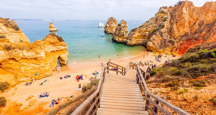 Holztreppe führt hinab zu einer malerischen Sandbucht, die von dramatischen goldenen Meeresfelsen und kristallklarem türkisfarbenem Wasser umrahmt wird, wo Strandbesucher sich entspannen und schwimmen