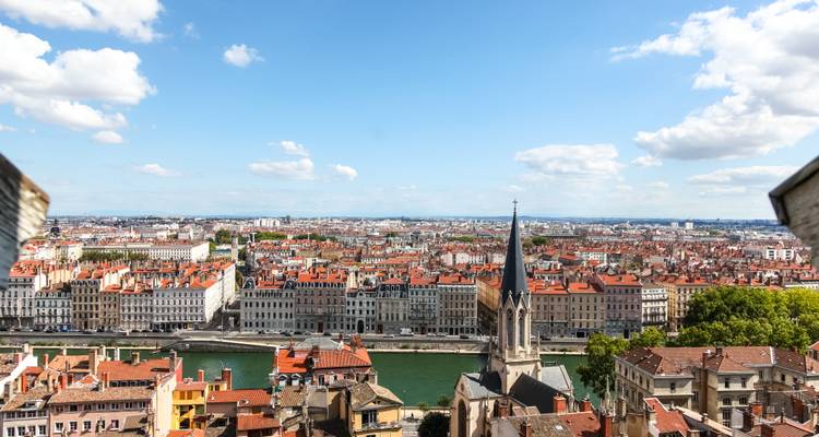 Erhöhte Panoramaaufnahme der Dächer von Lyon, eines Kirchturms und der Rhône unter einem strahlend blauen Himmel.