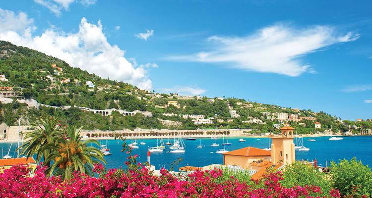 Vue encadrée de bougainvillées surplombant une baie d'un bleu éclatant parsemée de bateaux et de villas nichées à flanc de colline sur la Côte d'Azur.