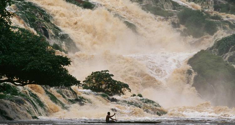 Un único remero en una canoa empequeñecido por una poderosa cascada de múltiples niveles que cae a través del terreno selvático.