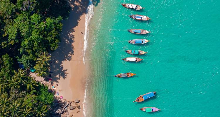 Vue aérienne par drone d'une baie turquoise avec des bateaux à longue queue traditionnels alignés le long d'une plage dorée.