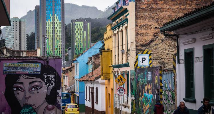 Calle llena de coloridos grafitis en el distrito histórico de Bogotá con torres modernas visibles a lo lejos.