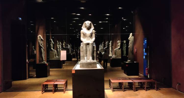Statue de pharaon en pierre exposée contre des murs en miroir dans une salle de musée sombre.