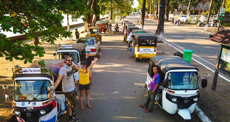 Reihe von Tuk-Tuks, die entlang der Straße am Fluss in Siem Reap geparkt sind, mit fröhlichen Reisenden, die winken.