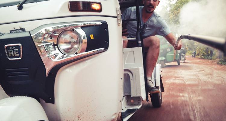 Aktionsaufnahme aus einem Tuk-Tuk-Rennen auf einer roten Schotterpiste, Reisender hält einen Selfiestab mit Staub in der Luft.
