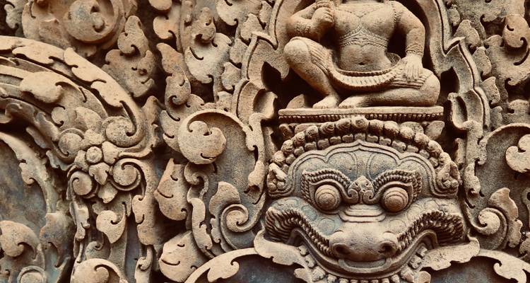 Detaillierte Nahaufnahme einer kunstvollen Sandsteinskulptur aus der Angkor-Ära mit einem mythischen Gesicht und einer Tänzerfigur.