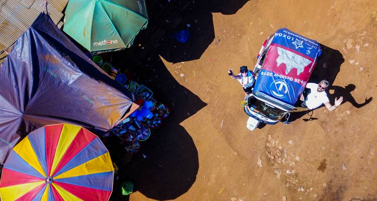 Drohnenaufnahme eines Tuk-Tuk-Rallye-Teams, das neben farbenfrohen Marktschirmen auf rotem Lehmboden winkt.