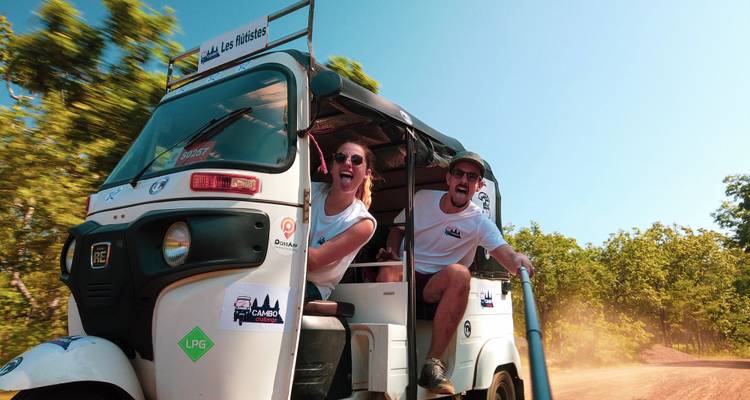 Zwei Reisende, die begeistert aus einem fahrenden Tuk-Tuk auf einer staubigen Waldstraße hängen.
