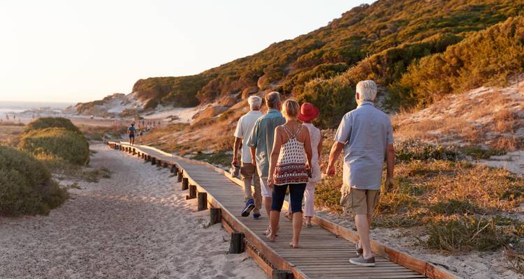 Groep oudere reizigers die langs een houten promenade wandelen aan een zandige kustduin bij zonsondergang