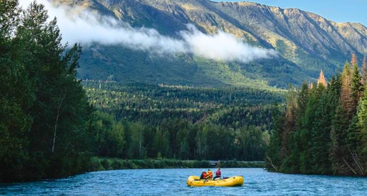 Deux aventuriers descendant en radeau un large fleuve de l'Alaska avec des pentes boisées et des montagnes au-delà.