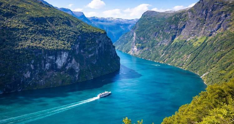 El espectacular Geirangerfjord de color turquesa serpentea entre montañas escarpadas y frondosas, con un crucero dejando su estela blanca.