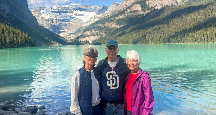 Trois voyageurs souriants se tiennent sur la rive rocheuse du lac Louise turquoise avec des pics couronnés de glaciers en arrière-plan.