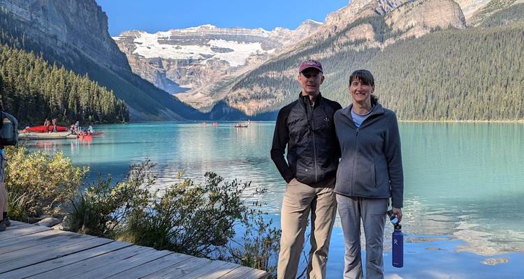 Un couple pose sur une passerelle en bois à côté des eaux turquoise du lac Louise avec des pics imposants en arrière-plan.