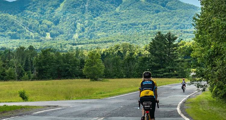 Cyclists pedal along a scenic country road framed by lush green mountains and forest in Vermont.
