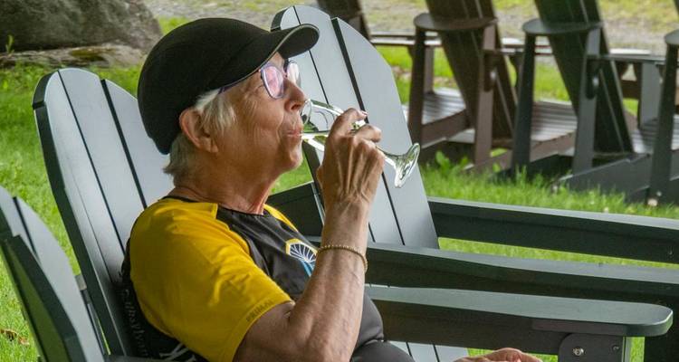Older woman relaxes in an Adirondack chair sipping a drink on a green lawn.
