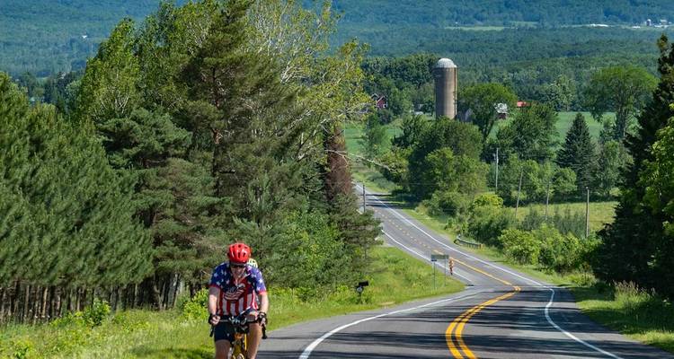 Cyclist wearing patriotic jersey climbs a rolling rural road past pine trees and a farm silo.
