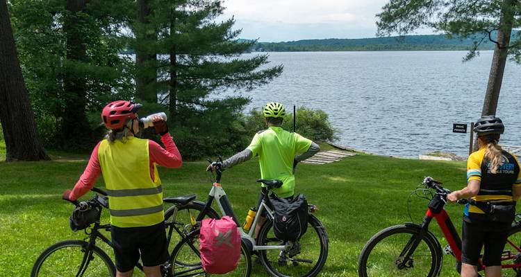 Three cyclists pause by a tranquil lakeshore, looking out over the water with their bikes.
