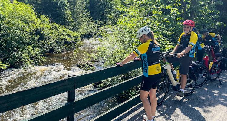Cyclists stop on a wooden bridge over a rushing river surrounded by lush greenery.
