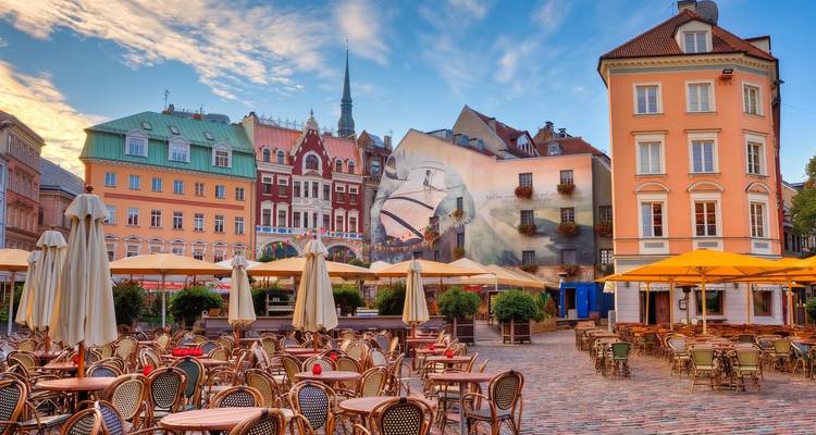 Charmanter Rigaer Platz, gesäumt von pastellfarbenen Gebäuden, Außencafés und leeren Korbstühlen unter dem Morgenhimmel.
