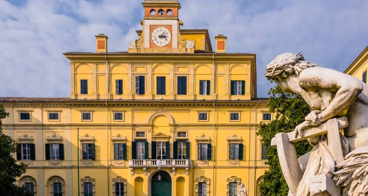 Yellow neoclassical palace with central clock and adjacent marble statue under bright morning light in Parma.