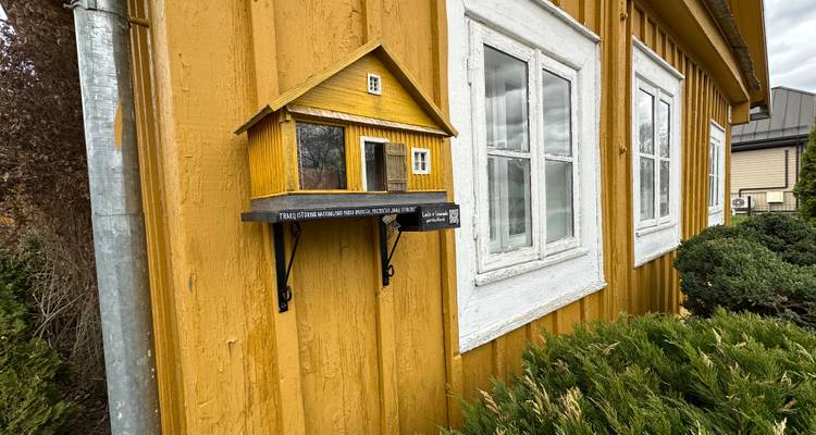 Una pequeña casita de pájaros de madera amarilla al estilo biblioteca montada en la pared de una cabaña de color mostaza.