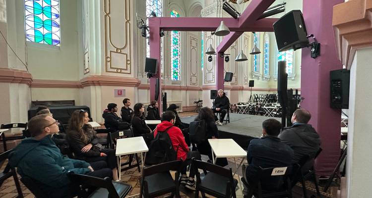 Grupo de turismo sentado dentro de un salón de iglesia convertido escuchando a un orador en un pequeño escenario.