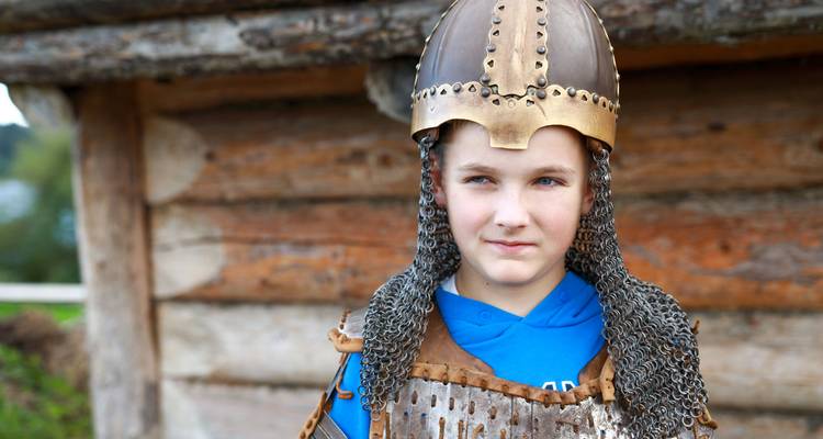 Un garçon portant un casque viking et une cotte de mailles se tient devant un bâtiment historique en bois.