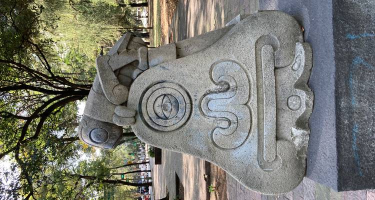 Aus Stein gehauerte Skulptur eines stilisierten Wesens, das auf einer abstrakten Basis in einem grünen Stadtpark angebracht ist