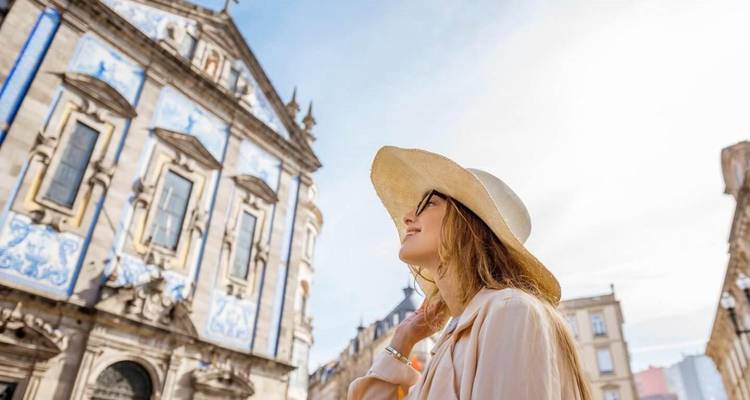 Femme souriante portant un chapeau à large bord regardant vers le haut la façade d'une église baroque aux carreaux bleus sous un ciel dégagé